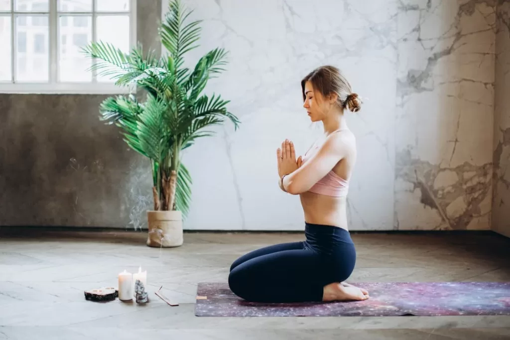 Mulher praticando yoga sozinha, sentada em posição de meditação, cercada por incenso e velas, ilustrando como fazer yoga sozinha.