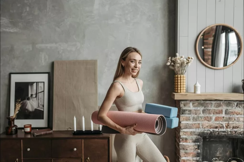 Uma mulher de pé sorrindo em uma sala com um tapete e um bloco de yoga, equipamentos essenciais para praticar yoga em casa.