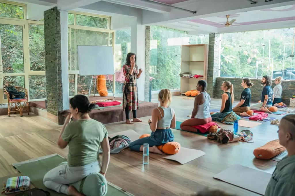 Um grupo de pessoas em um estúdio fazendo yoga sentadas em cima de bolsters de yoga ou almofadas cilíndricas relaxantes.