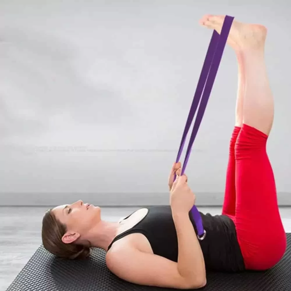 Uma mulher num estúdio de calça vermelha e regata preta com as costas apoiada no chão com um cinto de yoga roxo ajudando a ficar com as pernas levantadas.